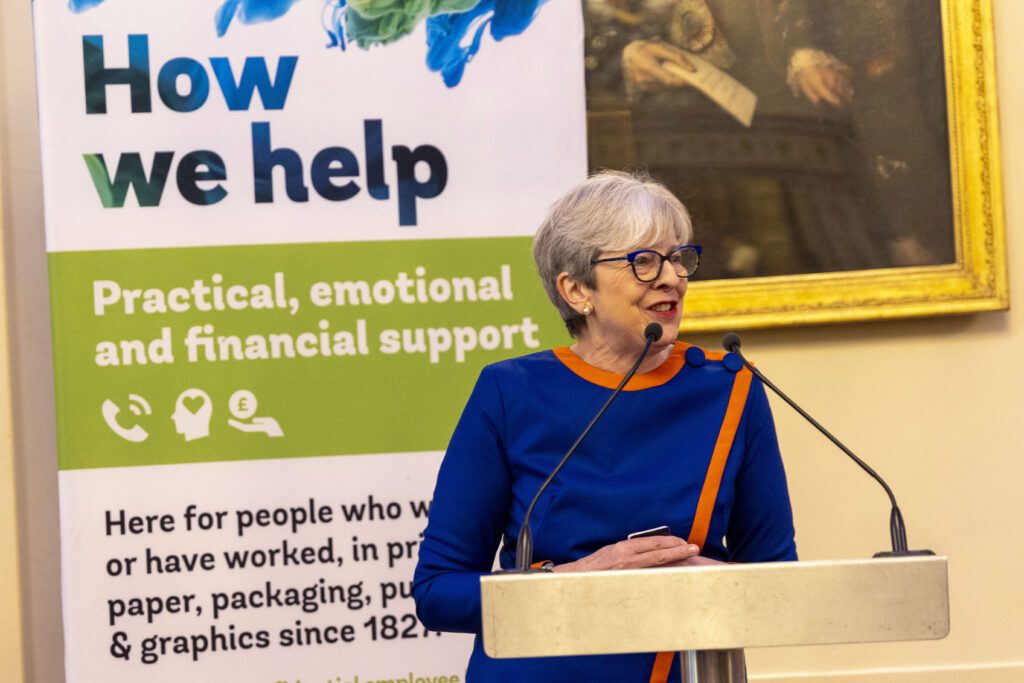 Baroness Theresa May speaking at podium at Printing Charity Annual Luncheon with charity banner displaying support services in background