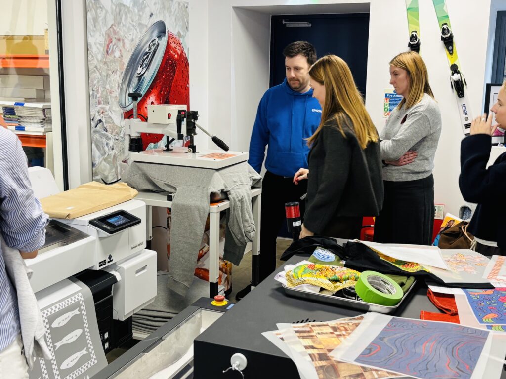 A group of students watches as a garment is loaded onto a digital direct-to-garment printer