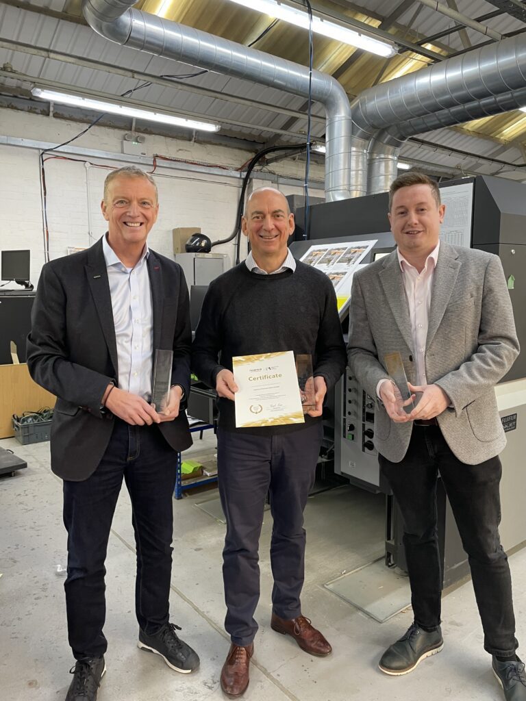 Three men standing in a print production facility holding certificates and trophies in front of a digital press.