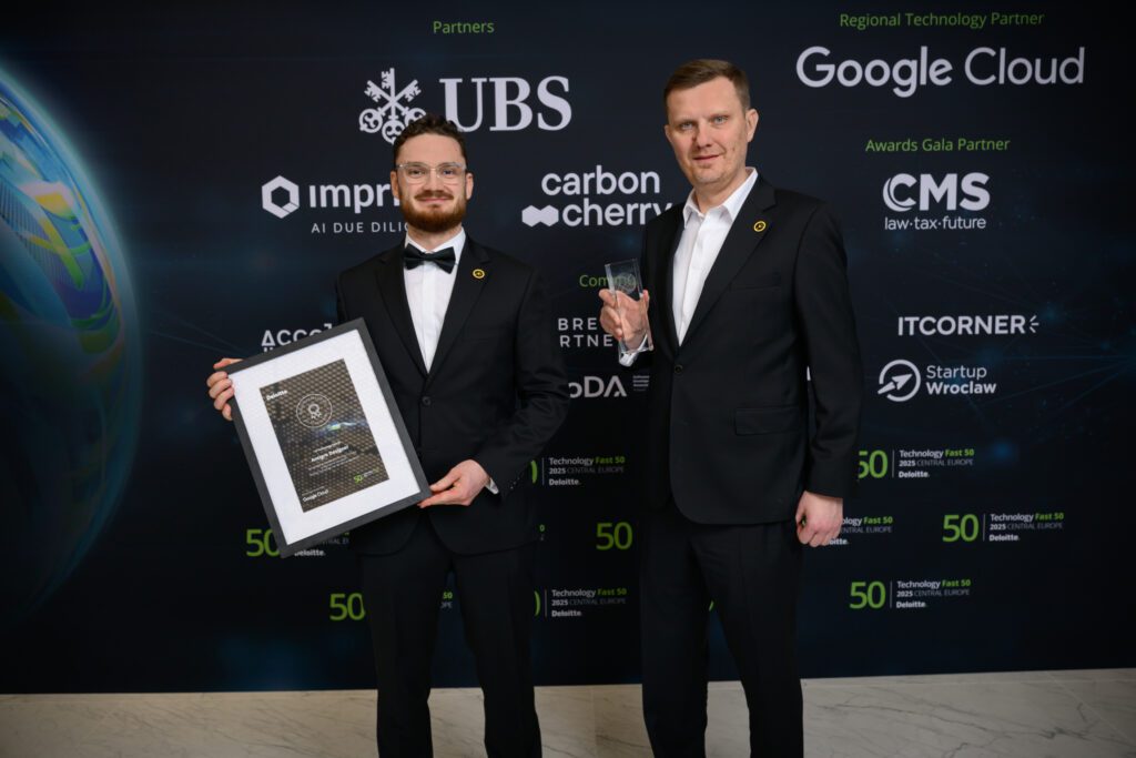 Wacław Mostowski and Marcin Majda standing in front of a media wall, holding a framed certificate and a glass trophy