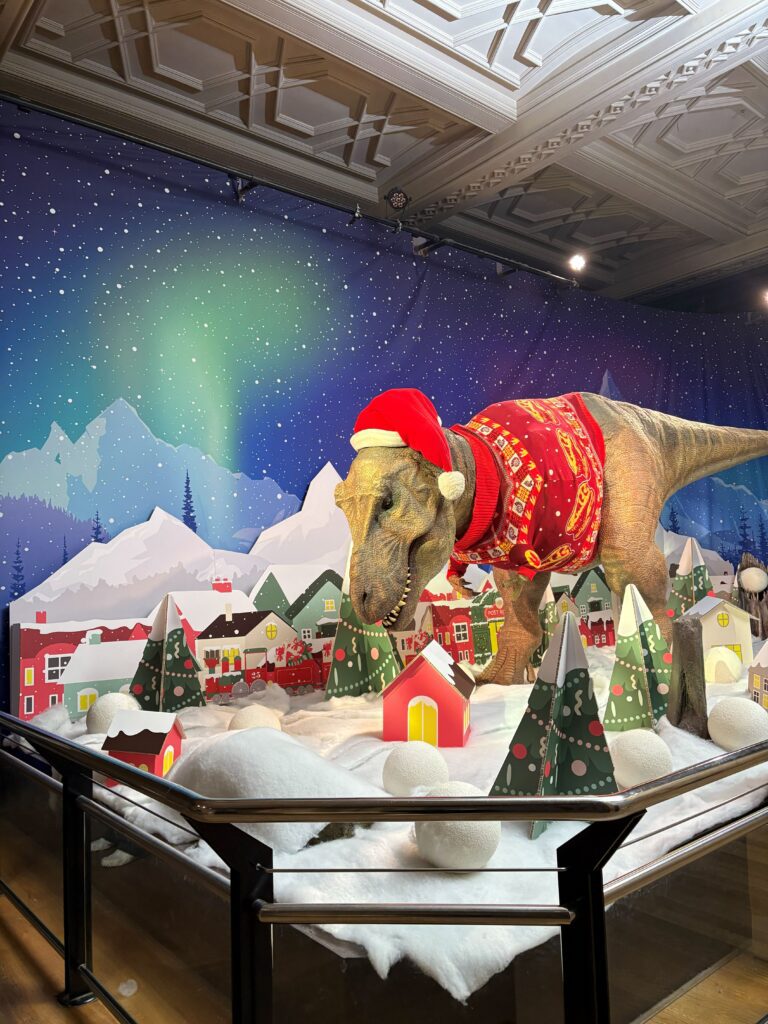 A creative festive display at the Natural History Museum featuring cardboard cut-outs decorated trees and houses in a snow scene with a T-rex in a Christmas jumper and hat