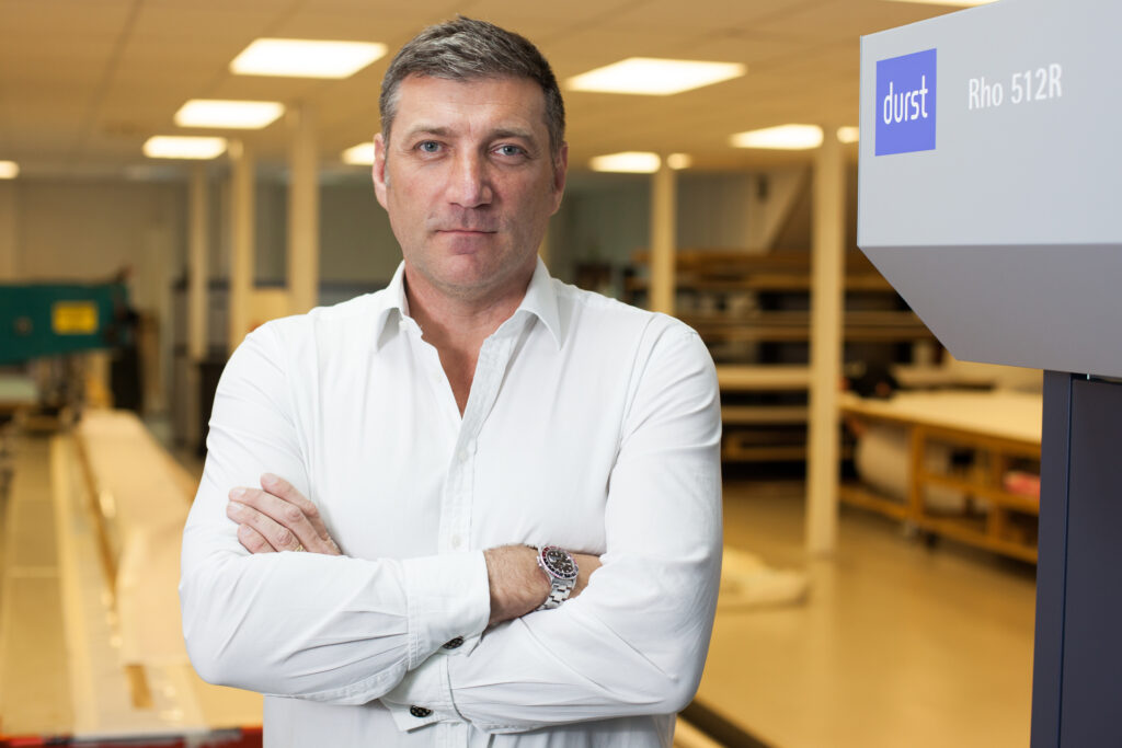 Matt Tydeman standing with arms crossed in a print production facility next to a Durst printer.