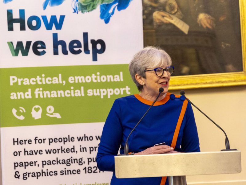 Baroness Theresa May speaking at podium at Printing Charity Annual Luncheon with charity banner displaying support services in background