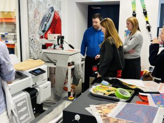 A group of students watches as a garment is loaded onto a digital direct-to-garment printer