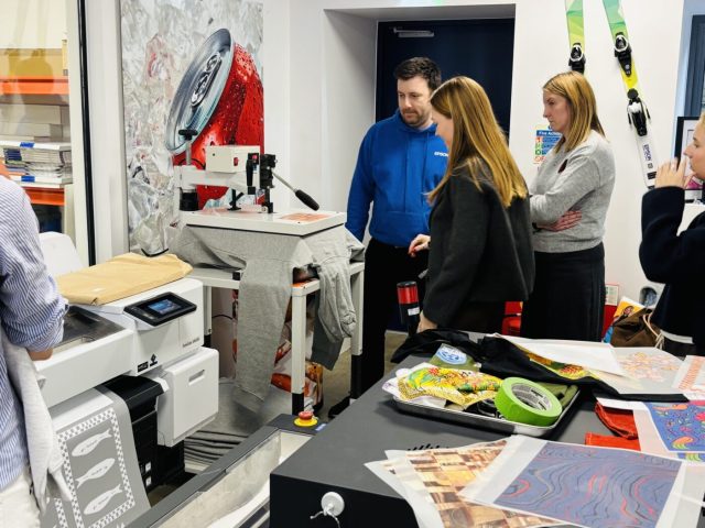 A group of students watches as a garment is loaded onto a digital direct-to-garment printer