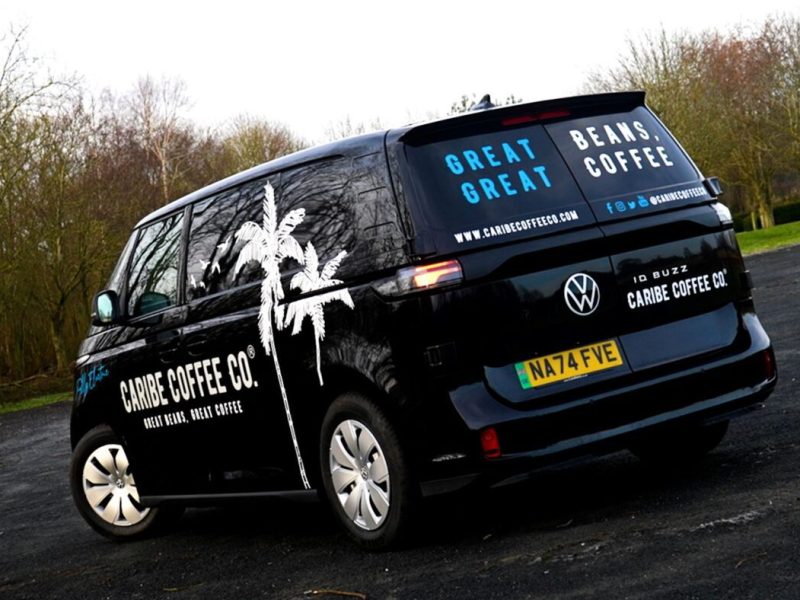 A black VW ID Buzz van with white "Caribe Coffee Co." branding and palm tree graphics parked outdoors