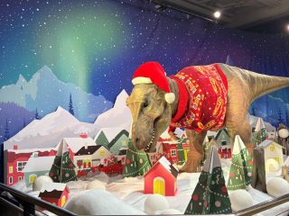 A creative festive display at the Natural History Museum featuring cardboard cut-outs decorated trees and houses in a snow scene with a T-rex in a Christmas jumper and hat