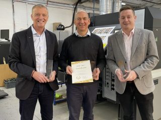 Three men standing in a print production facility holding certificates and trophies in front of a digital press.