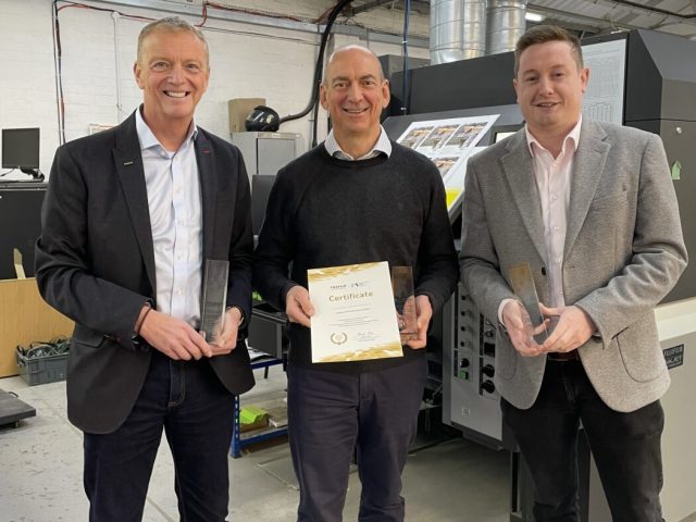 Three men standing in a print production facility holding certificates and trophies in front of a digital press.