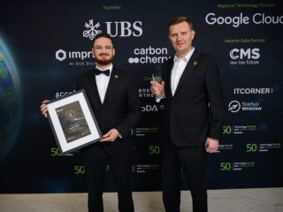 Wacław Mostowski and Marcin Majda standing in front of a media wall, holding a framed certificate and a glass trophy